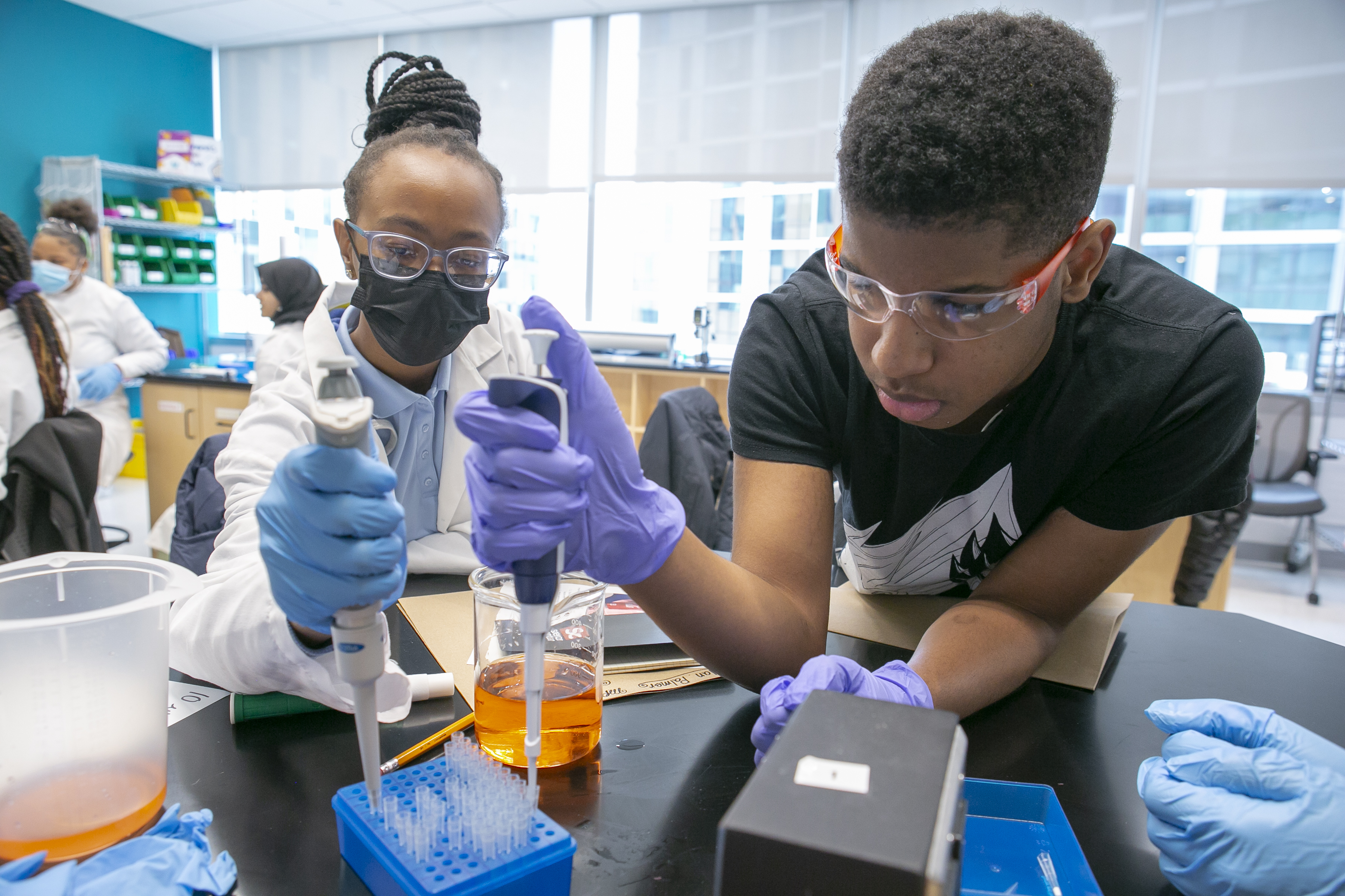 Two younger students wearing lab coats and gloves working together on a project in a lab.